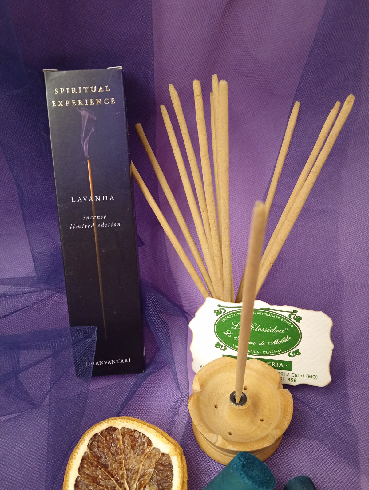 Astuccio di Incenso Spiritual Experience Lavanda Dhanvantari in verticale con mazzo di bastoncini e portaincenso in legno chiaro, fetta di arancia essiccata, legnetti e pietra azzurra su sfondo con velo viola, Erboristeria La Clessidra Carpi