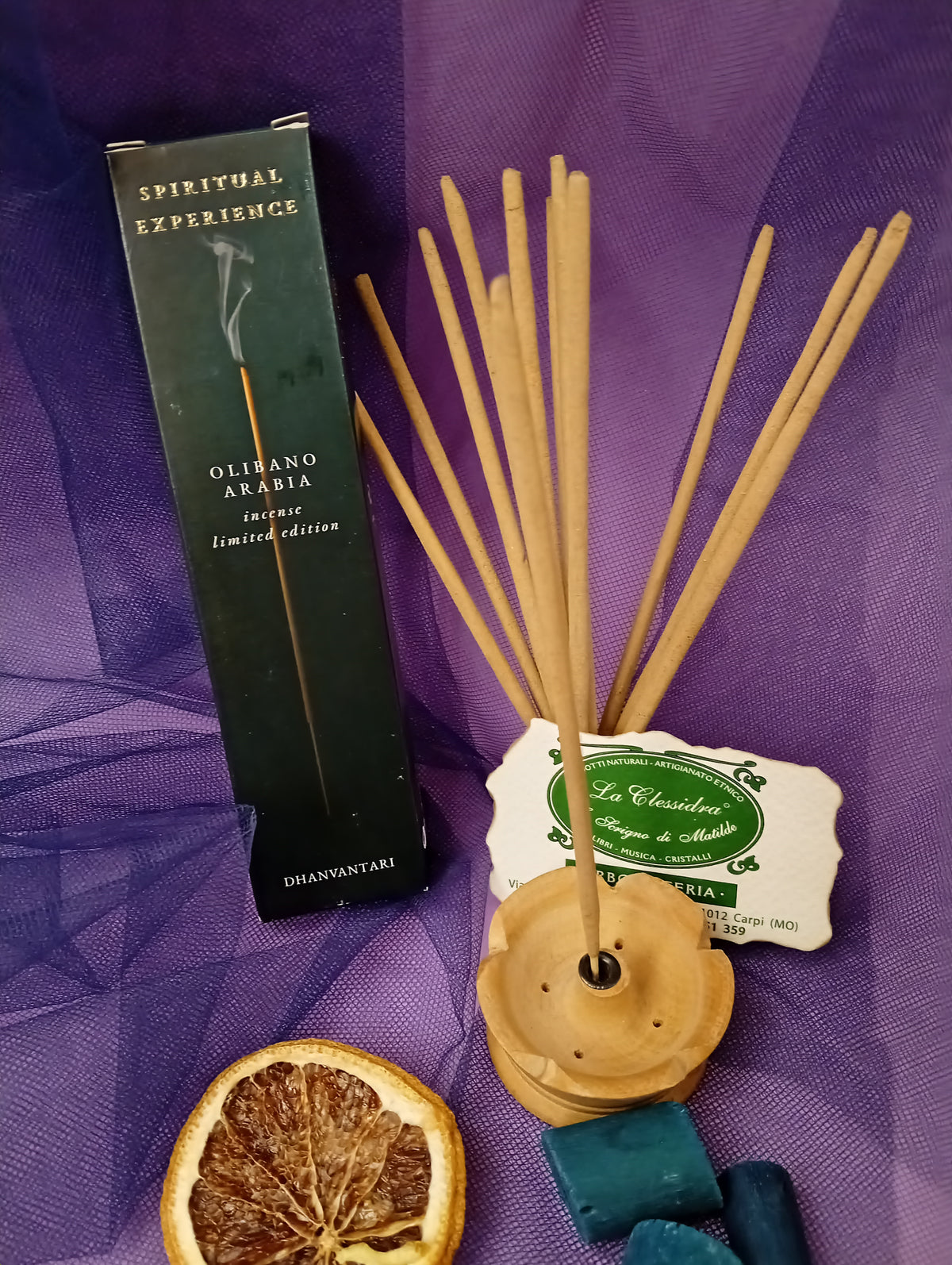 Astuccio di Incenso Spiritual Experience Olibano Arabia Dhanvantari in verticale con mazzo di bastoncini e portaincenso in legno chiaro, fetta di arancia essiccata, legnetti e pietra azzurra su sfondo con velo viola, Erboristeria La Clessidra Carpi