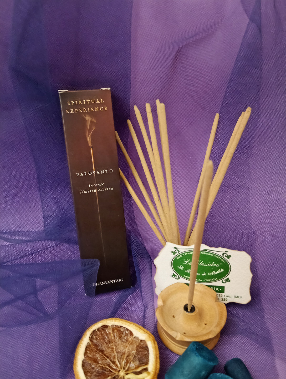 Astuccio di Incenso Spiritual Experience Palo Santo Dhanvantari in verticale con mazzo di bastoncini e portaincenso in legno chiaro, fetta di arancia essiccata, legnetti e pietra azzurra su sfondo con velo viola, Erboristeria La Clessidra Carpi