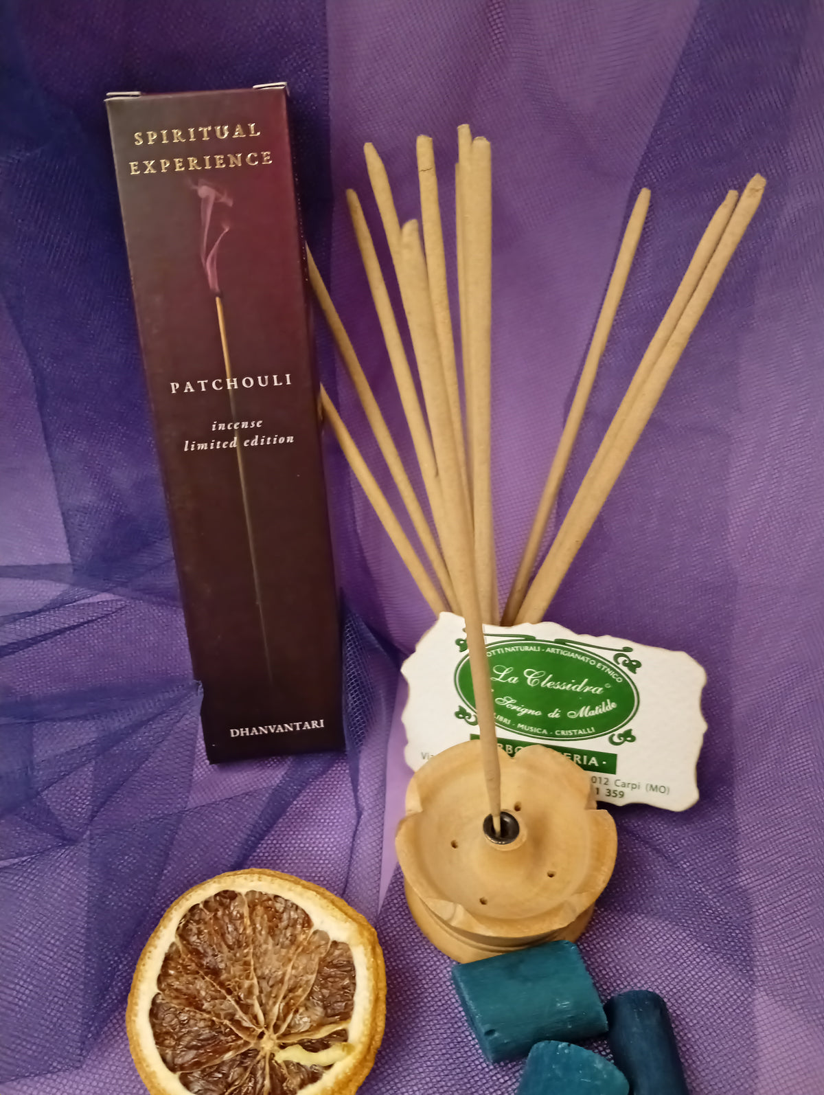 Astuccio di Incenso Spiritual Experience Patchouli Dhanvantari in verticale esposizione di bastoncini e portaincenso in legno chiaro, fetta di arancia essiccata, legnetti e pietra azzurra su sfondo con velo viola, Erboristeria La Clessidra Carpi