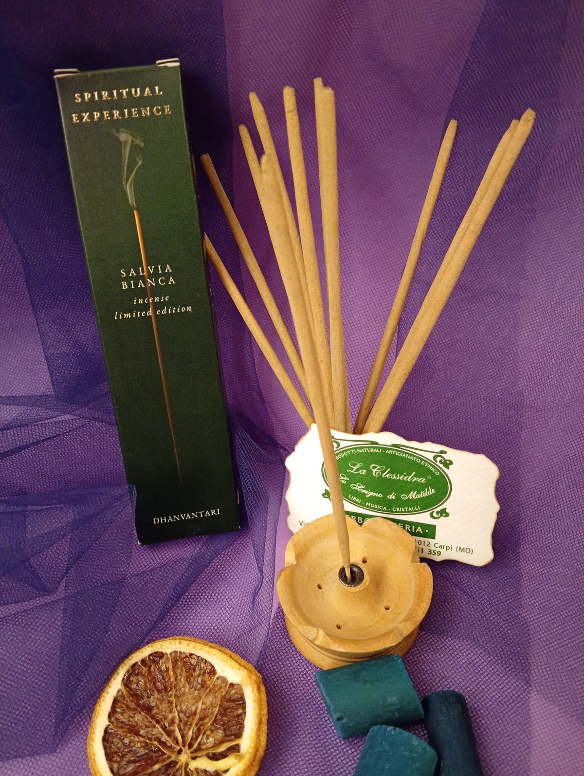Astuccio di Incenso Spiritual Experience Salvia Bianca Dhanvantari in verticale con mazzo di bastoncini e portaincenso in legno chiaro, fetta di arancia essiccata, legnetti e pietra azzurra su sfondo con velo viola, Erboristeria La Clessidra Carpi