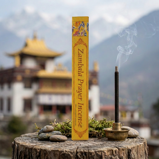 Confezione di incenso tibetano Zambala Prayer Himalayan Arts su un tronco con sassi e muschio, accanto a un bastoncino acceso, con tempio tibetano e montagne sullo sfondo